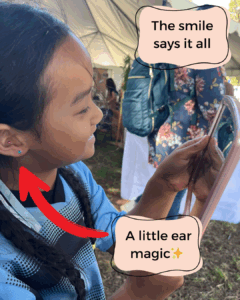 A girl smiling happily at their reflection in a mirror after an auriculotherapy session, with visible ear seeds in place on their outer ear. Little Sage Acupuncture in Long Beach